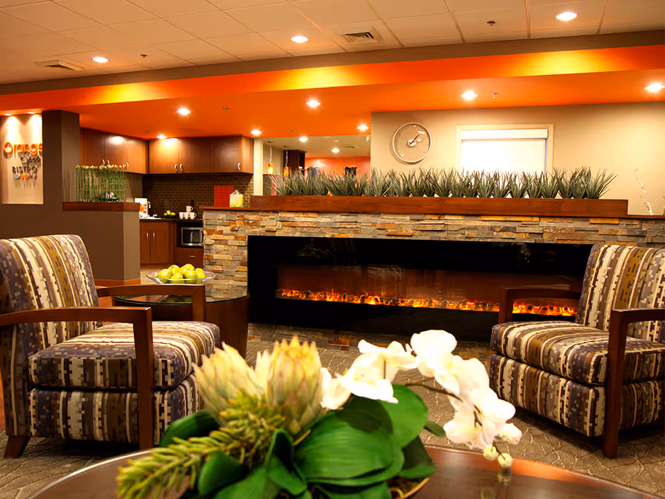 Warm lobby lounge with fireplace, two patterned chairs, plants, and a coffee table.