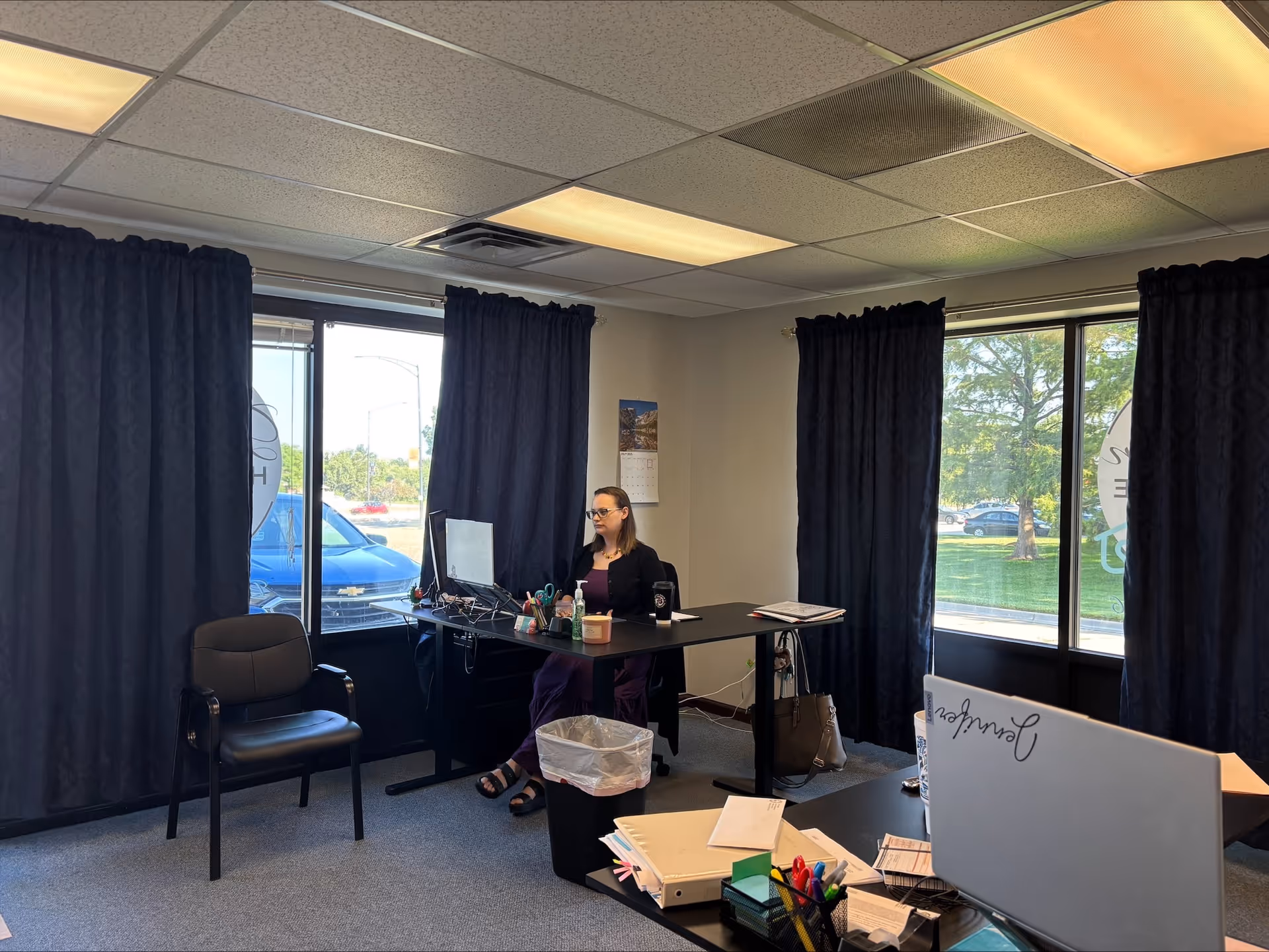 Woman seated at a desk with computer in a small office with blue curtains.