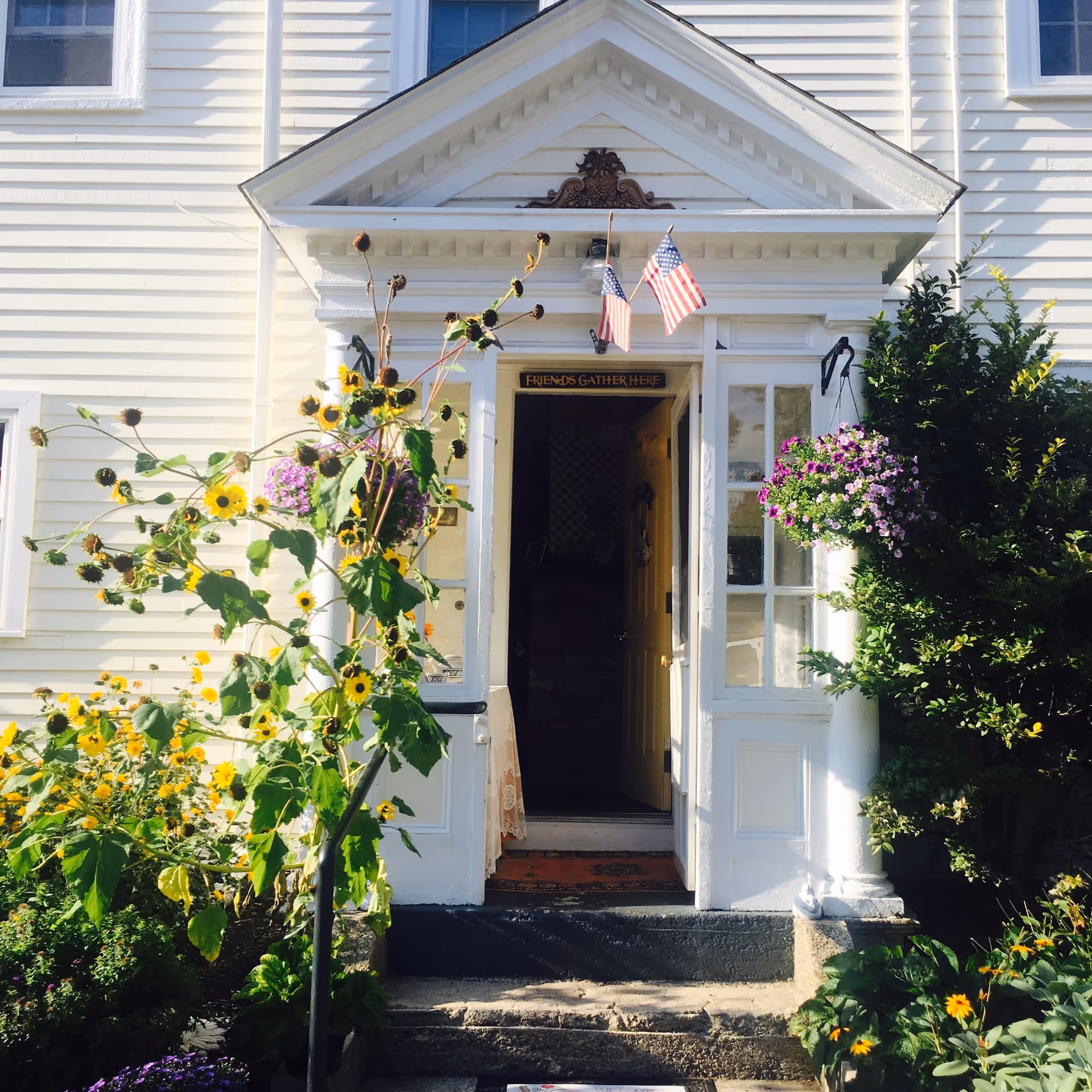 White house-style building entrance with blooming flowers, sunflowers, and American flags.