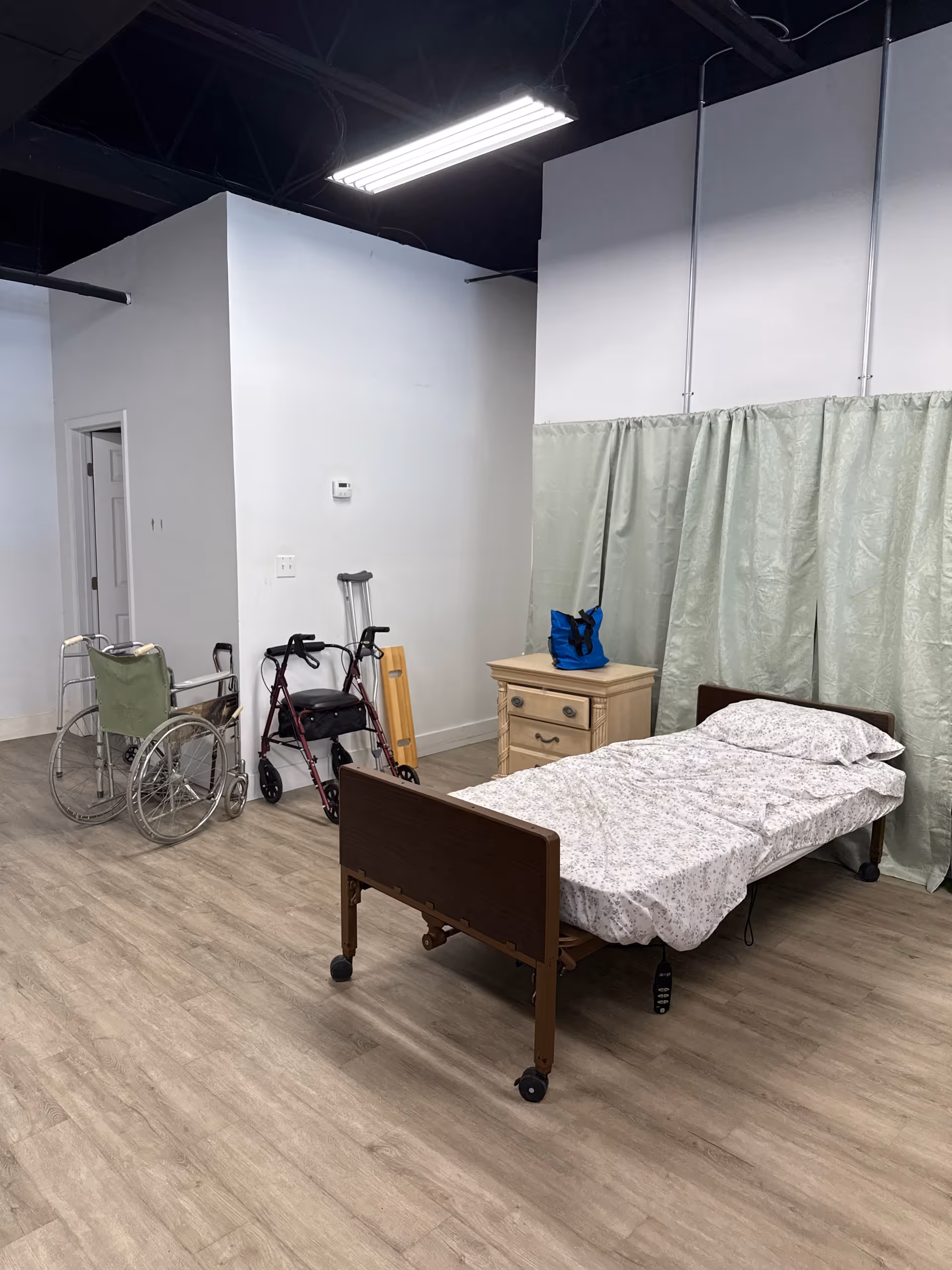 Hospital-style bedroom with bed, privacy curtain, wheelchair, walker, and dresser.