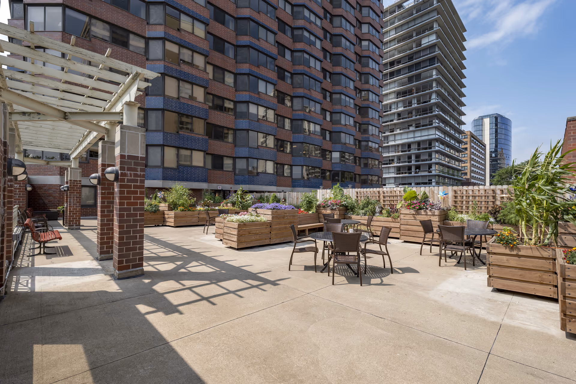 Outdoor patio area with several round tables and chairs surrounded by large wooden planters filled with various flowers and plants. The patio is adjacent to a multi-story brick and blue building, with other tall buildings visible in the background under a clear blue sky.