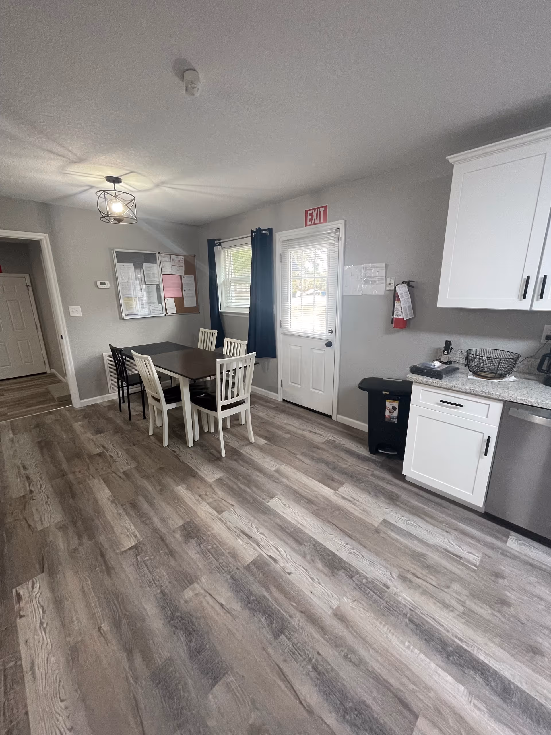 A bright dining area with a dark table, white chairs, and a kitchenette along the wall.