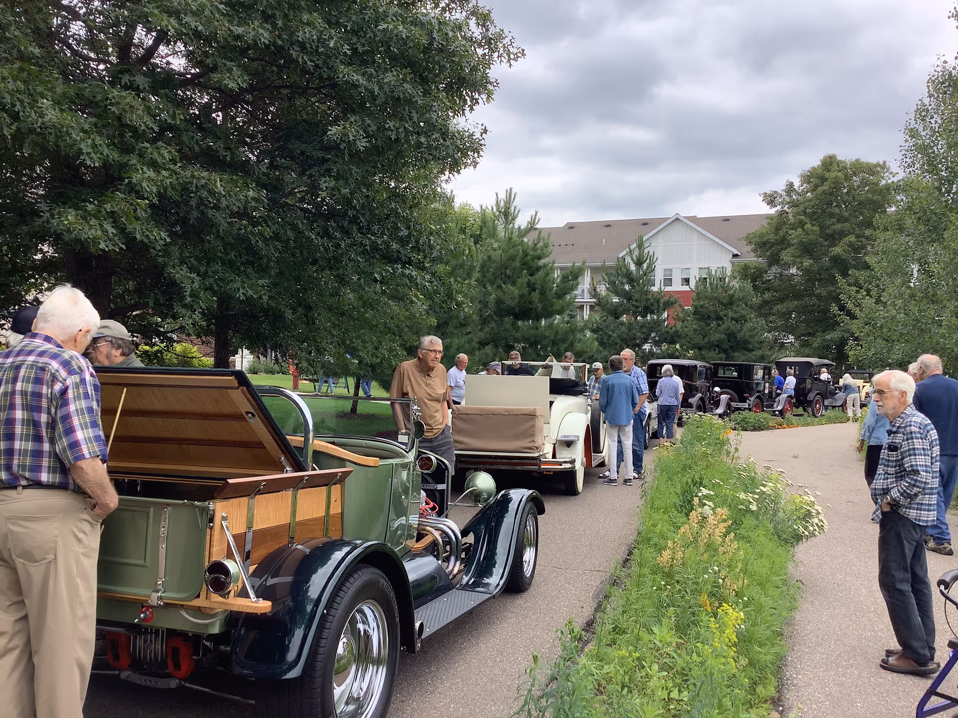 A group of elderly people gathered outdoors along a paved pathway with several vintage cars parked on one side. The scene is set in a green, leafy area with trees and shrubs, and a large building is visible in the background under a cloudy sky.
