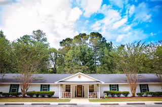 Front exterior view of a single-story white building with a gray roof, surrounded by trees and bushes under a partly cloudy blue sky. The building has a central entrance with a small covered porch and symmetrical windows on either side.