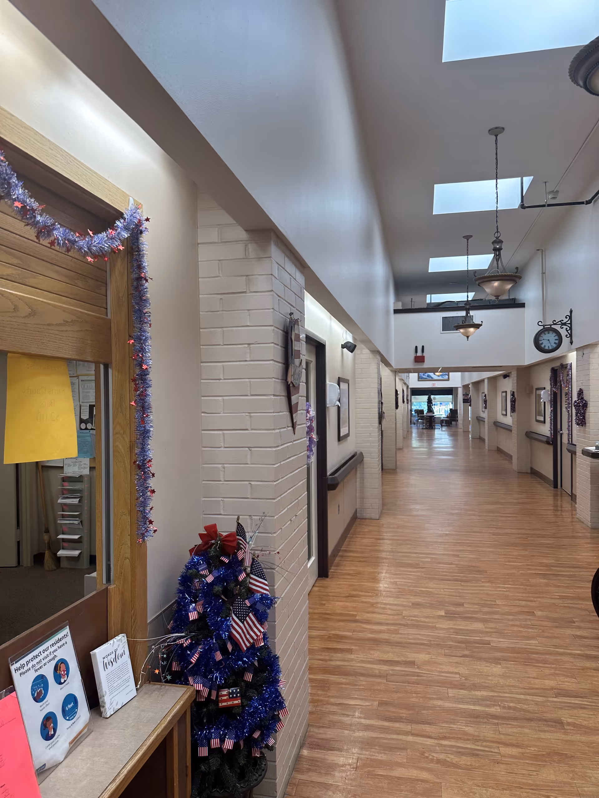 A long, well-lit hallway inside a senior living facility with wooden flooring and white brick walls. The hallway is decorated with patriotic-themed garlands and small American flags. There are several doors along the hallway, a clock mounted on the wall, and hanging light fixtures from the ceiling. Skylights provide additional natural light.