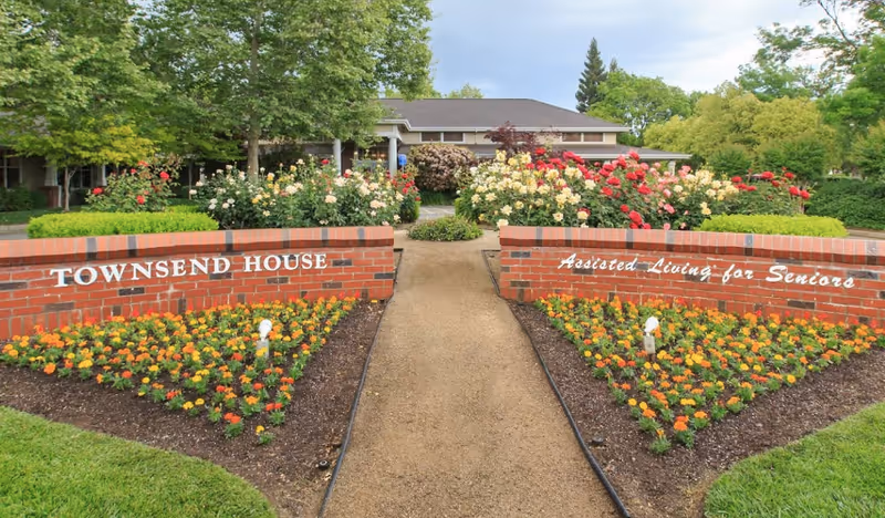 Entrance pathway leading to a building surrounded by well-maintained flower beds with orange and yellow flowers, and bushes with red and white roses. Two brick walls flank the pathway, one with the text 'TOWNSEND HOUSE' and the other with 'Assisted Living for Seniors'. Trees and greenery are visible in the background under a partly cloudy sky.