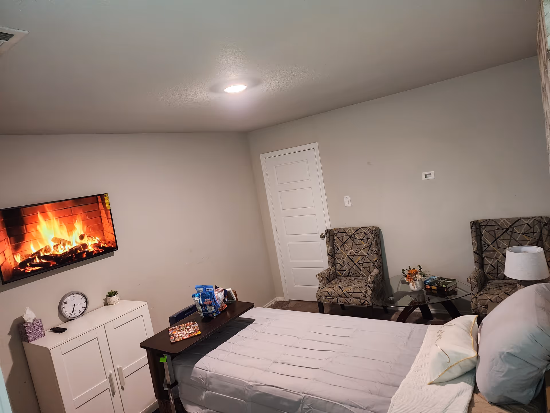 Cozy bedroom with TV fireplace, two chairs, and a bed.