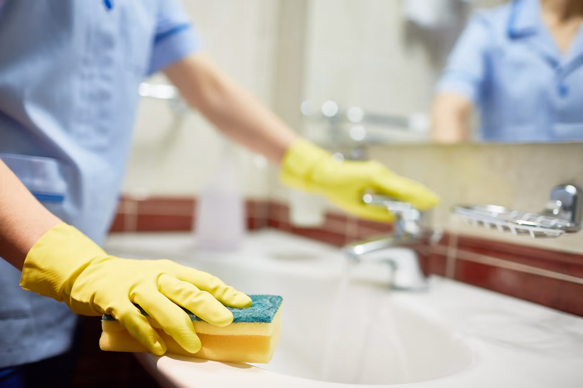 A caregiver wearing yellow gloves cleans a bathroom sink with a sponge near a mirror.