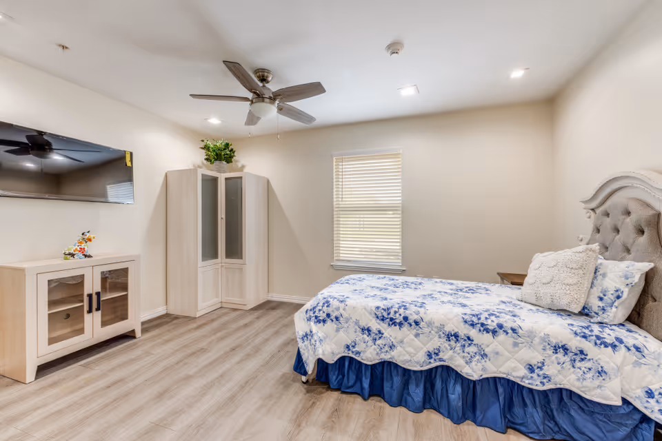 A furnished bedroom in a senior living facility with a queen bed and light wood furniture.