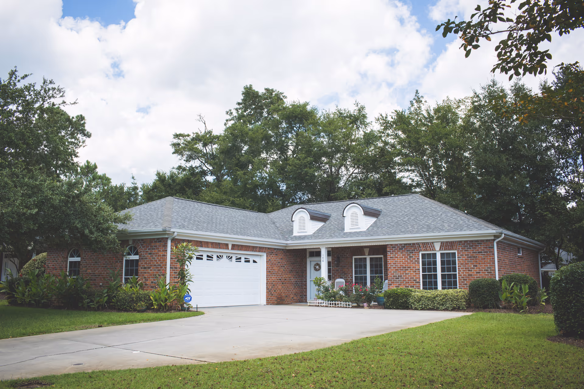 Single-story brick house with a two-car garage, driveway, and landscaped front yard surrounded by trees.