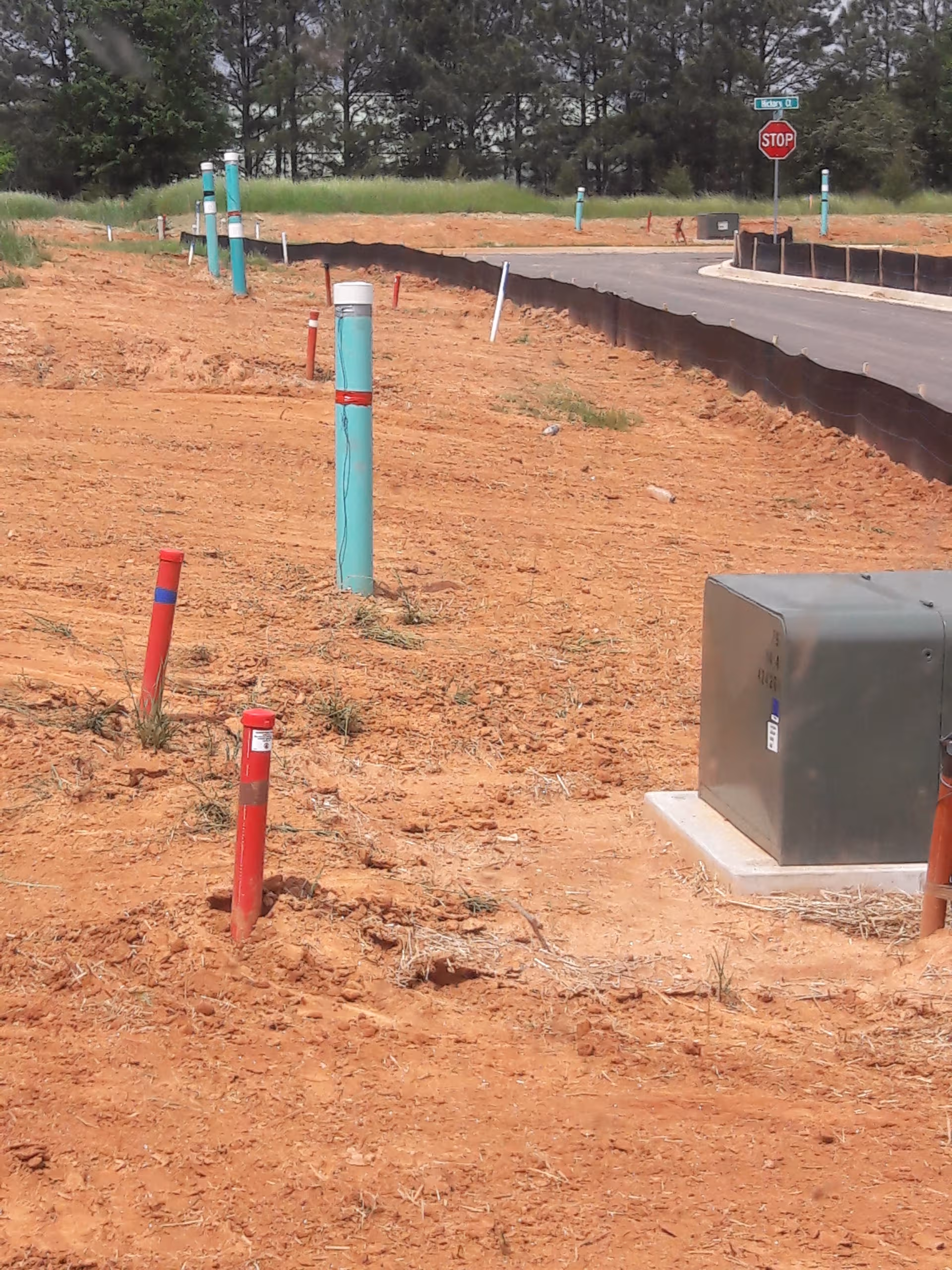 Construction site with exposed pipes, dirt, and a metal utility box beside a road.