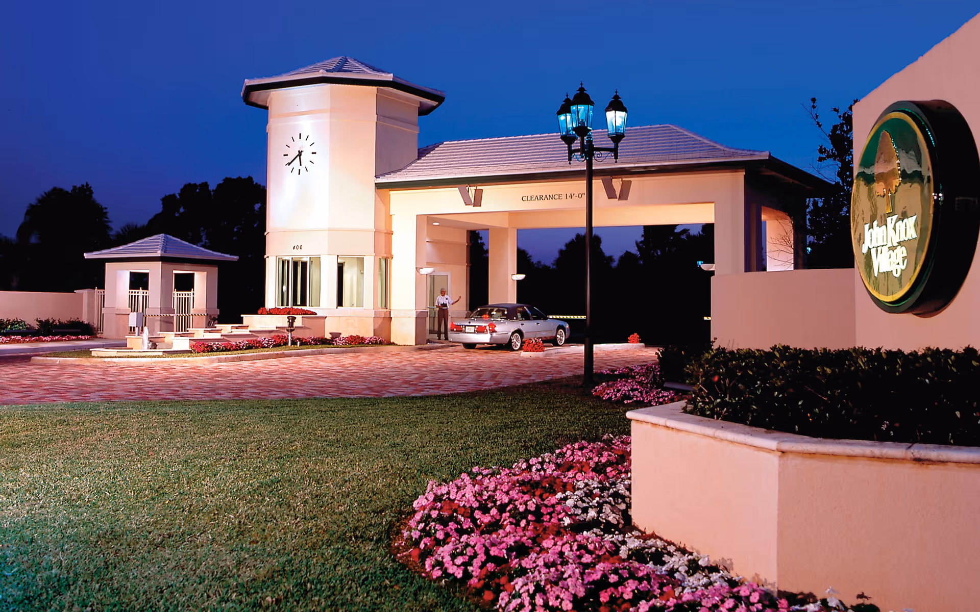 Entrance gate of John Knox Village at dusk with a clock tower, a car passing through, well-maintained landscaping with green grass and pink flowers, and a sign displaying the facility name.