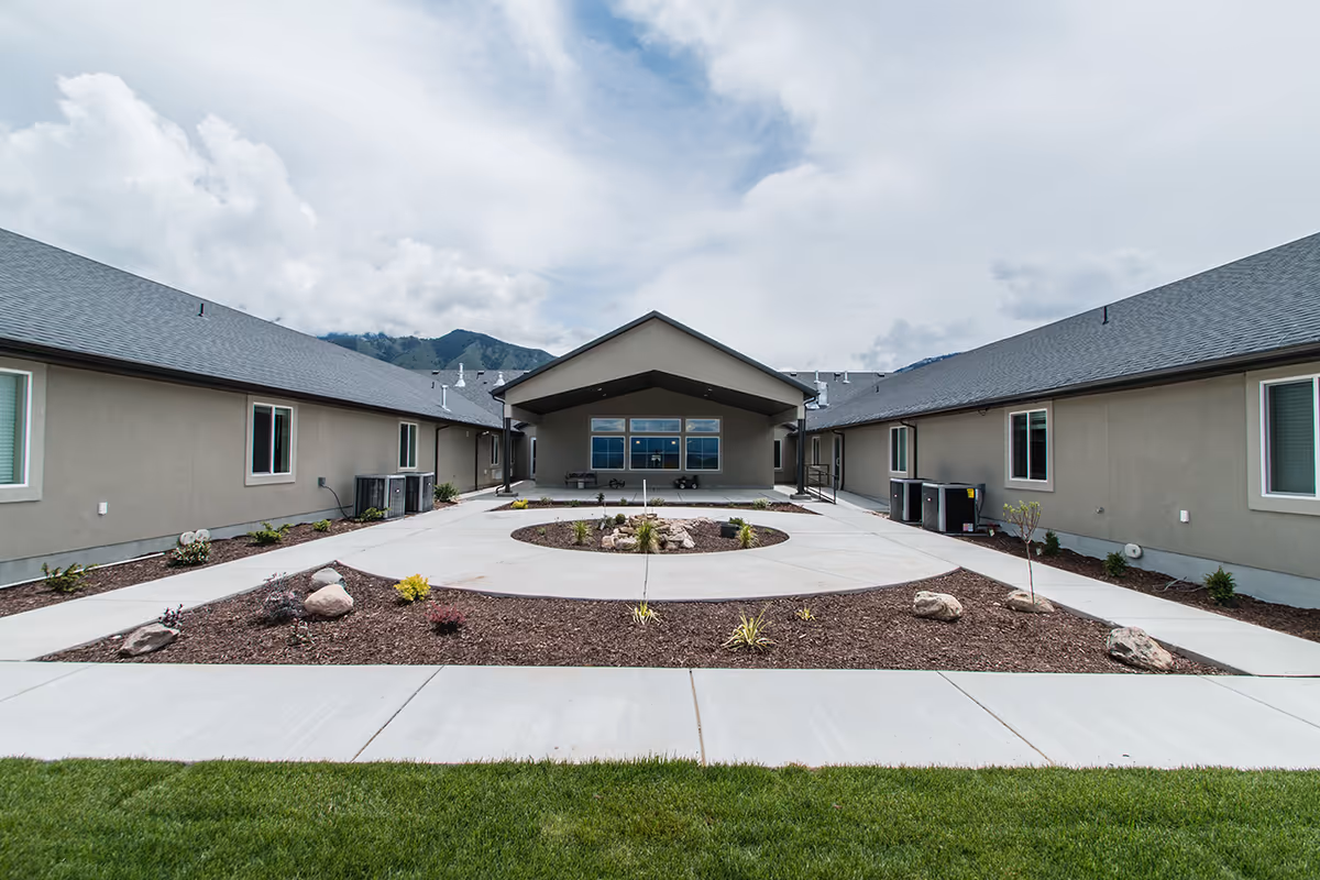 Courtyard and covered entrance of a single-story assisted living building with walkways, minimal landscaping, and mountains in the background.