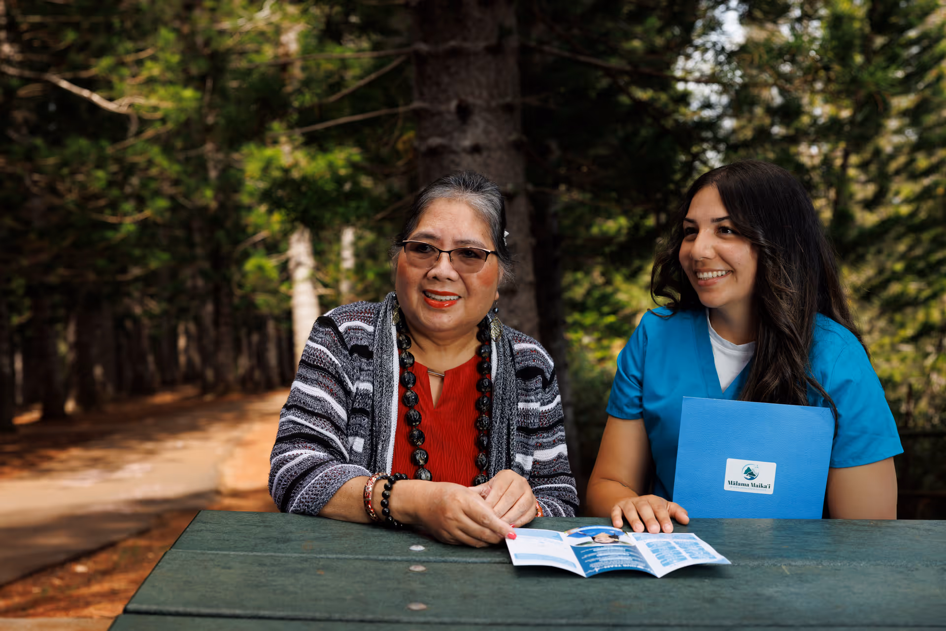 Two women sit at an outdoor picnic table smiling, one caregiver in blue scrubs.