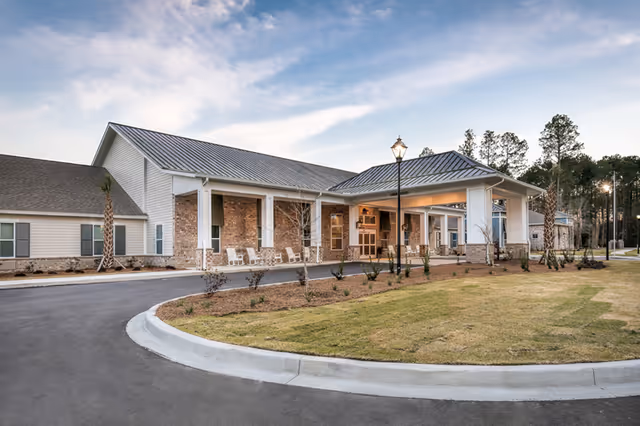 Exterior view of Sprenger Health Care Bluffton building with a covered entrance, brick and siding facade, a curved driveway, landscaped lawn, and trees in the background under a partly cloudy sky.