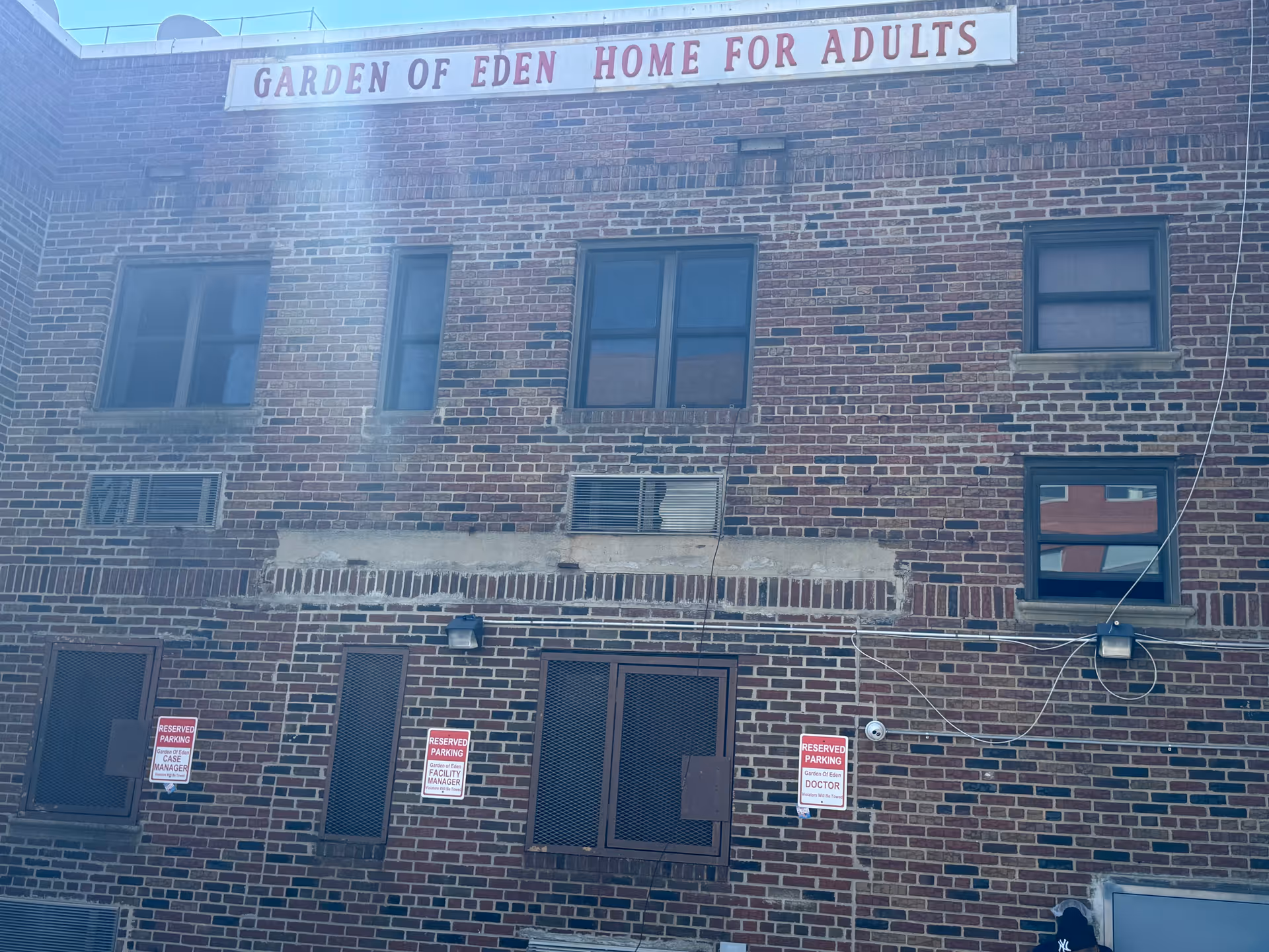 Brick facade of the Garden of Eden Home for Adults building with windows, a rooftop sign, and reserved parking signs on the lower level.