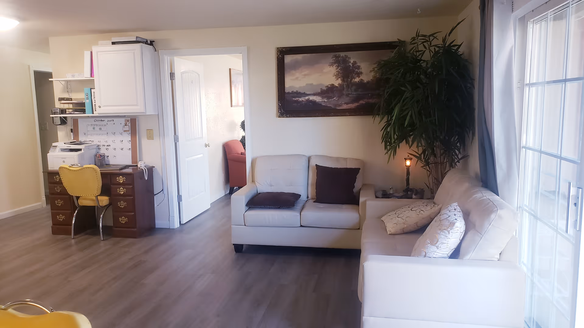 A cozy living room in a senior facility with white sofas, a desk, and plants.