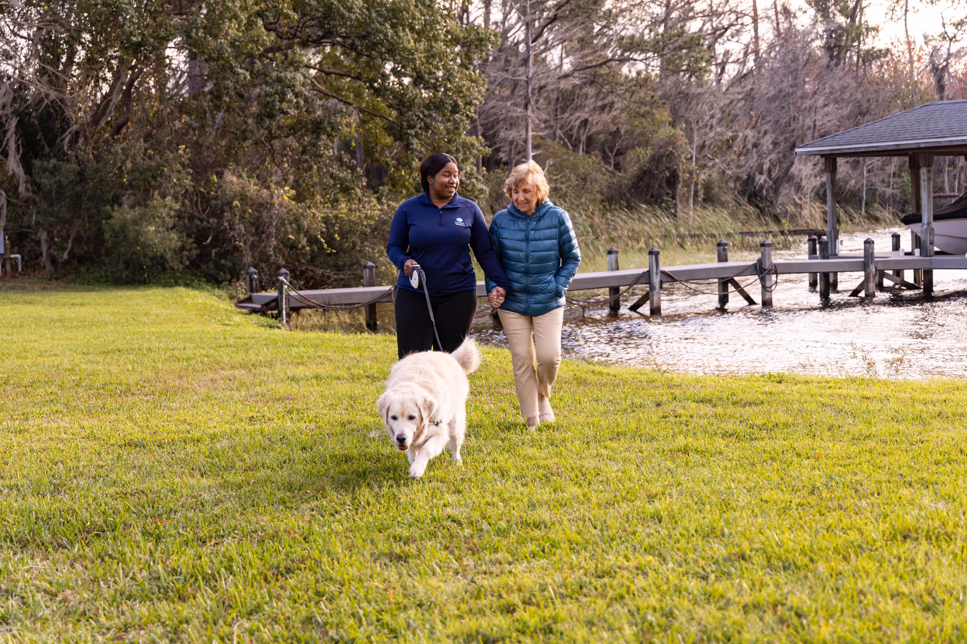 Two people walk a dog on a grassy lakeside area near a dock.