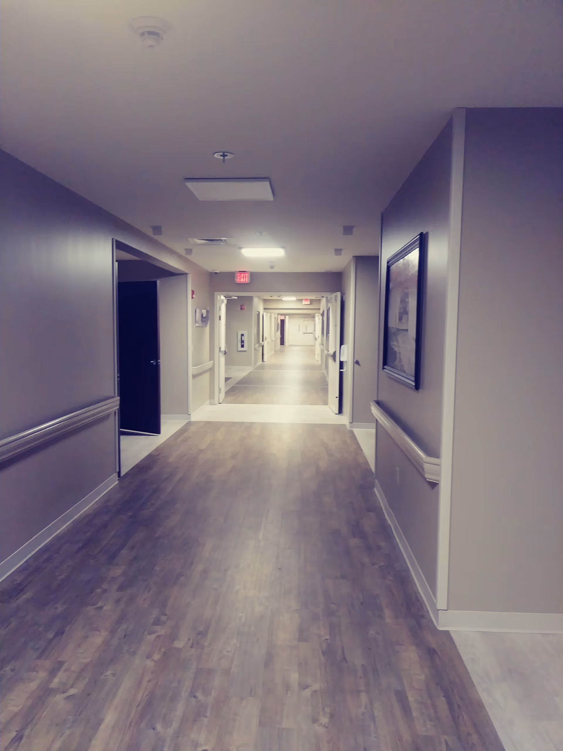 Long, well-lit hallway with doors, artwork, and wood-look flooring in a care facility.