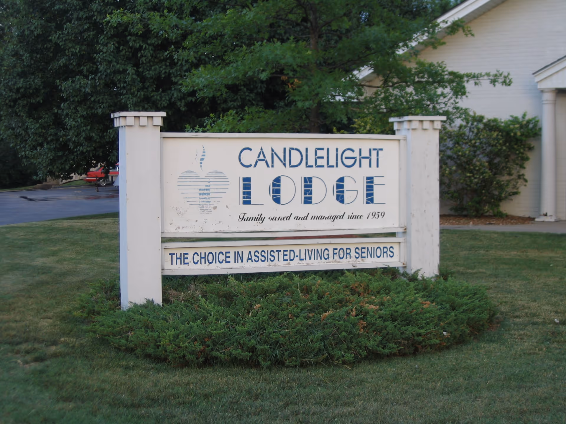 Outdoor sign for Candlelight Lodge, an assisted living facility for seniors, situated on a grassy area with bushes and trees in the background.