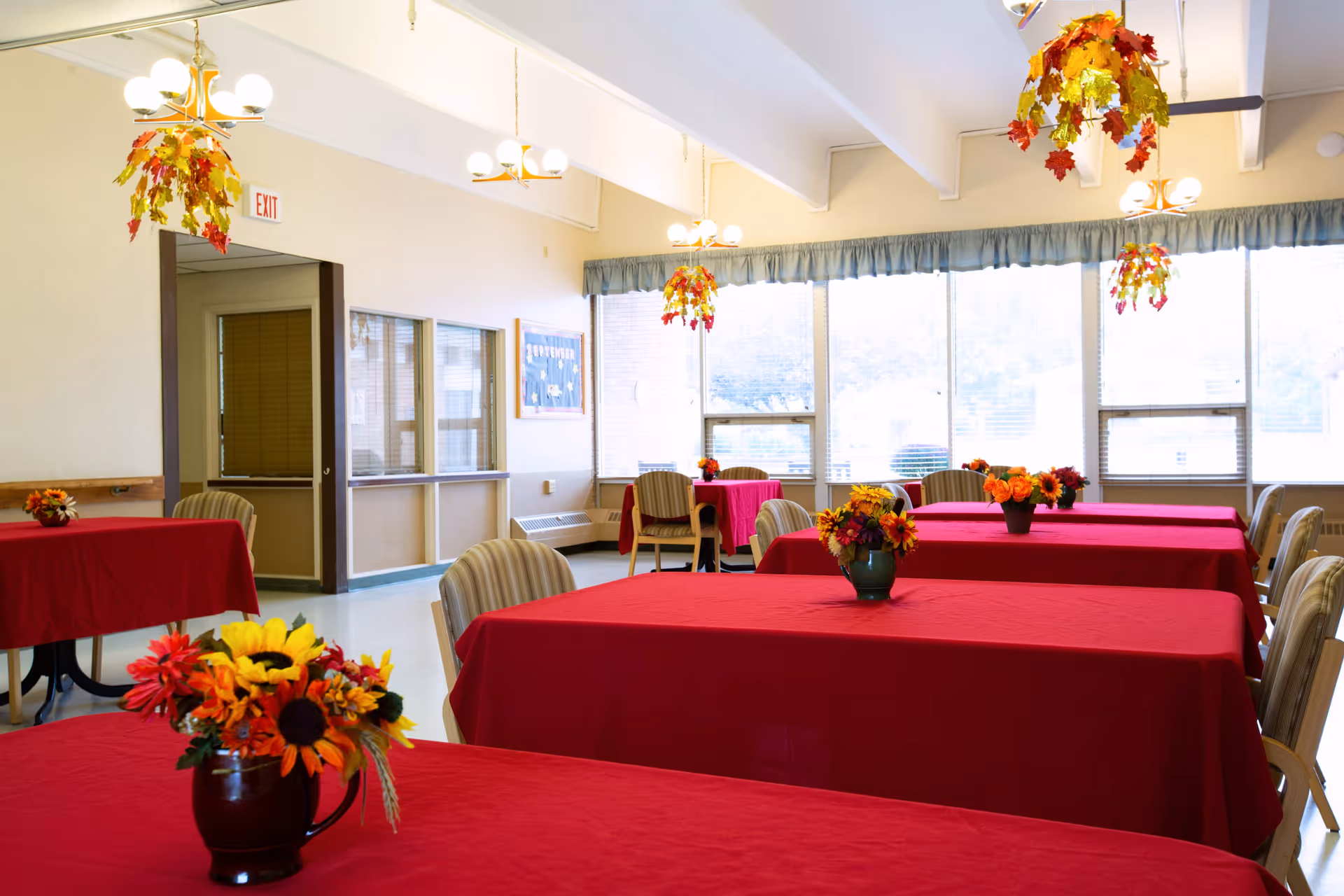 A bright dining room with several tables covered in red tablecloths, each adorned with a vase of colorful artificial flowers. The room has large windows with blue curtains, letting in natural light. Ceiling lights are decorated with autumn leaf arrangements, and there are beige cushioned chairs around the tables.