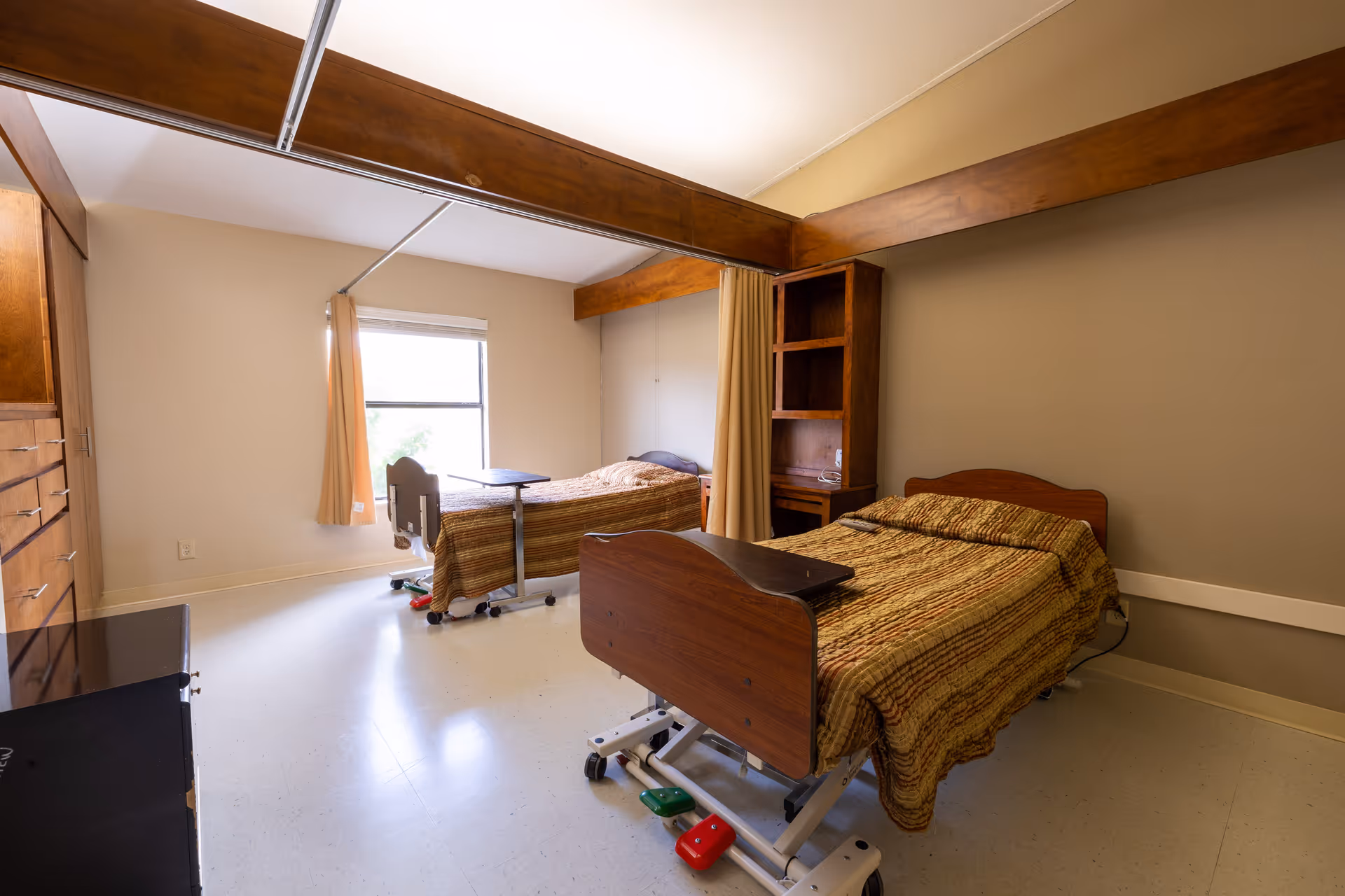 A nursing facility room with two hospital beds separated by a curtain. Each bed has a brown striped blanket and an overbed table. There is a window with beige curtains letting in natural light, wooden cabinetry, and a built-in shelving unit on the wall.