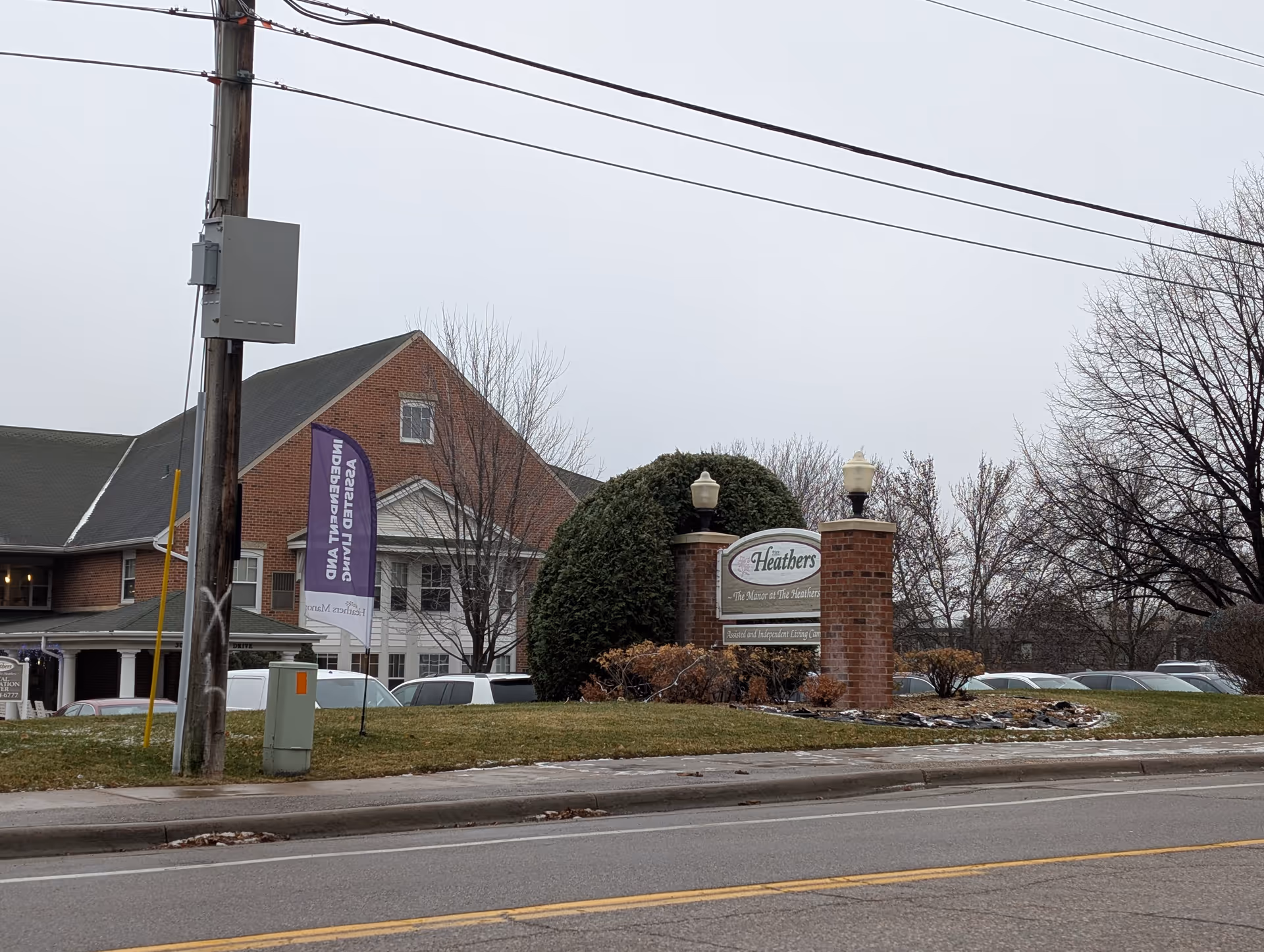 Exterior view of Heathers Manor, a brick building with a green lawn and a sign in front that reads 'Heathers - The Manor at The Heathers - Assisted and Independent Living Community'. Several cars are parked behind the sign, and there are leafless trees and a cloudy sky in the background.