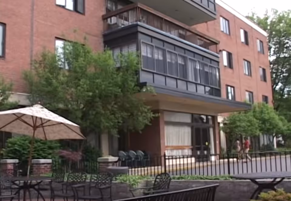 Entrance and outdoor patio area of a multi-story brick senior living building with tables, an umbrella, and landscaping.