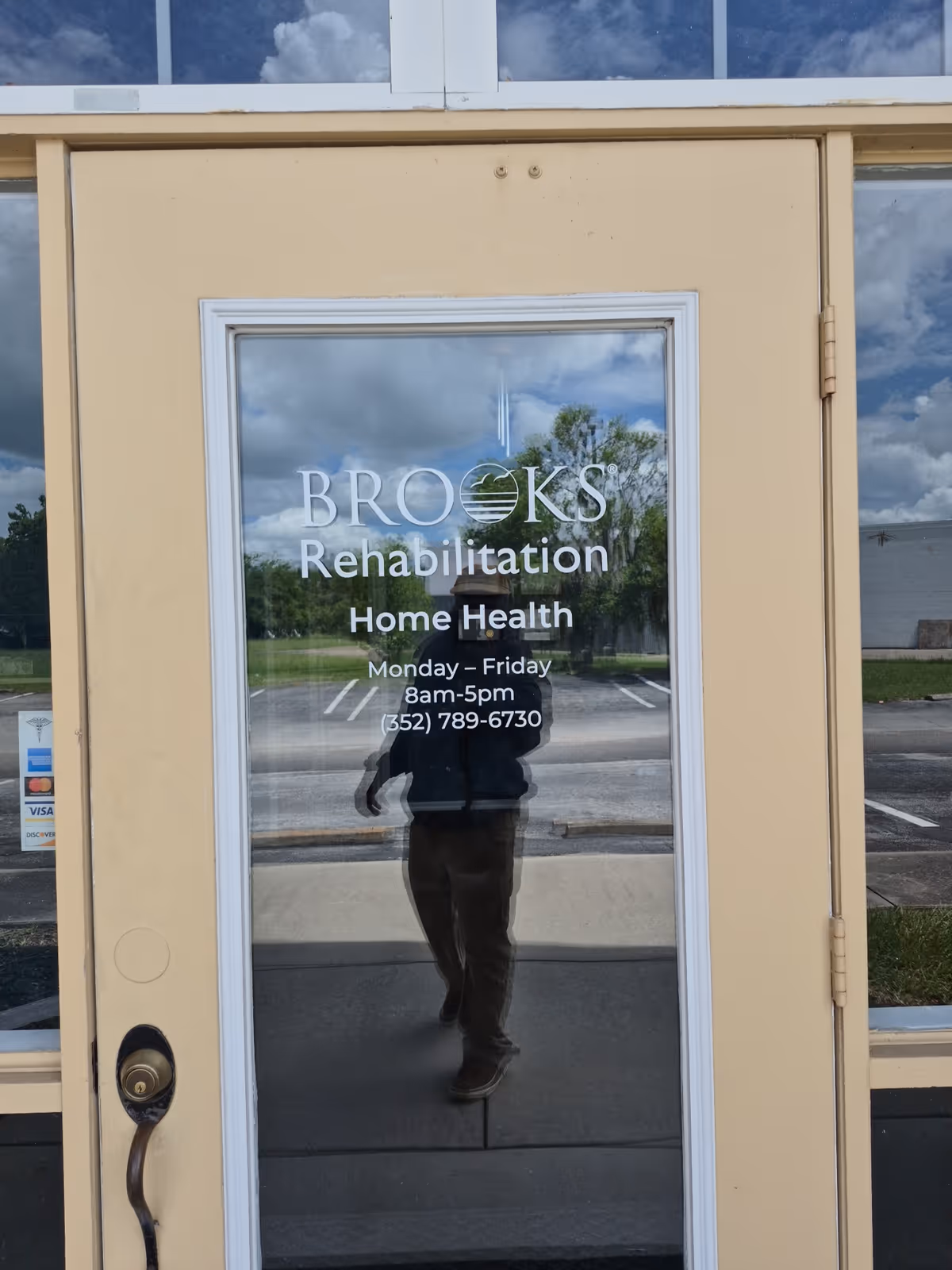 Glass front door displays Brooks Rehabilitation Home Health signage with a reflected person taking a photo.