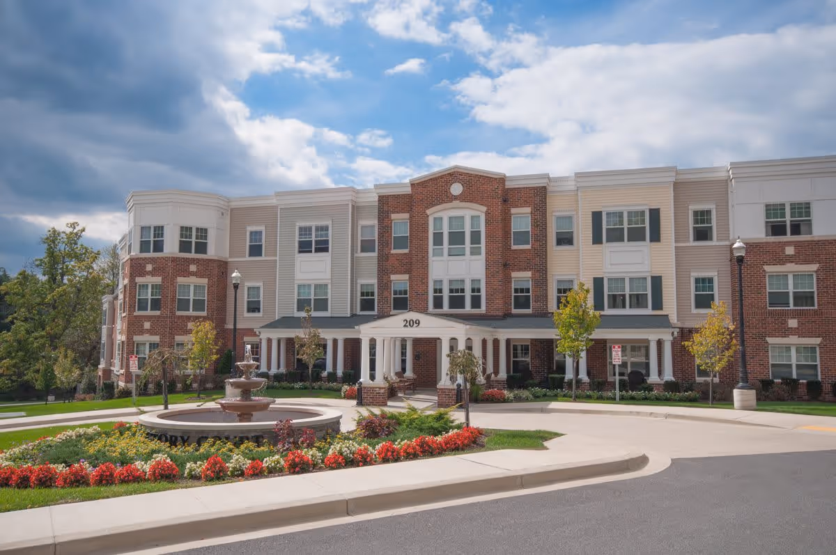 Front exterior view of Victory Court Senior Apartments, a three-story building with a combination of brick and light-colored siding. The entrance features white columns and a covered porch with the number 209 above it. In front of the building is a circular driveway with a tiered water fountain surrounded by colorful flowers and landscaped greenery. The sky is partly cloudy.