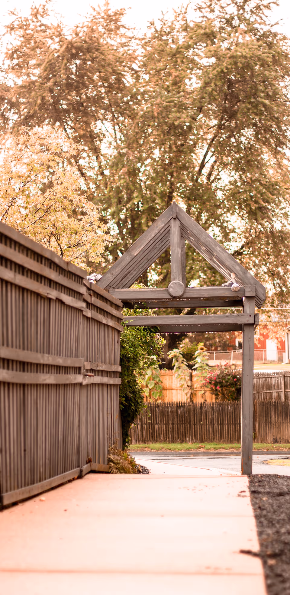 Wooden archway over a paved outdoor path beside a fence.