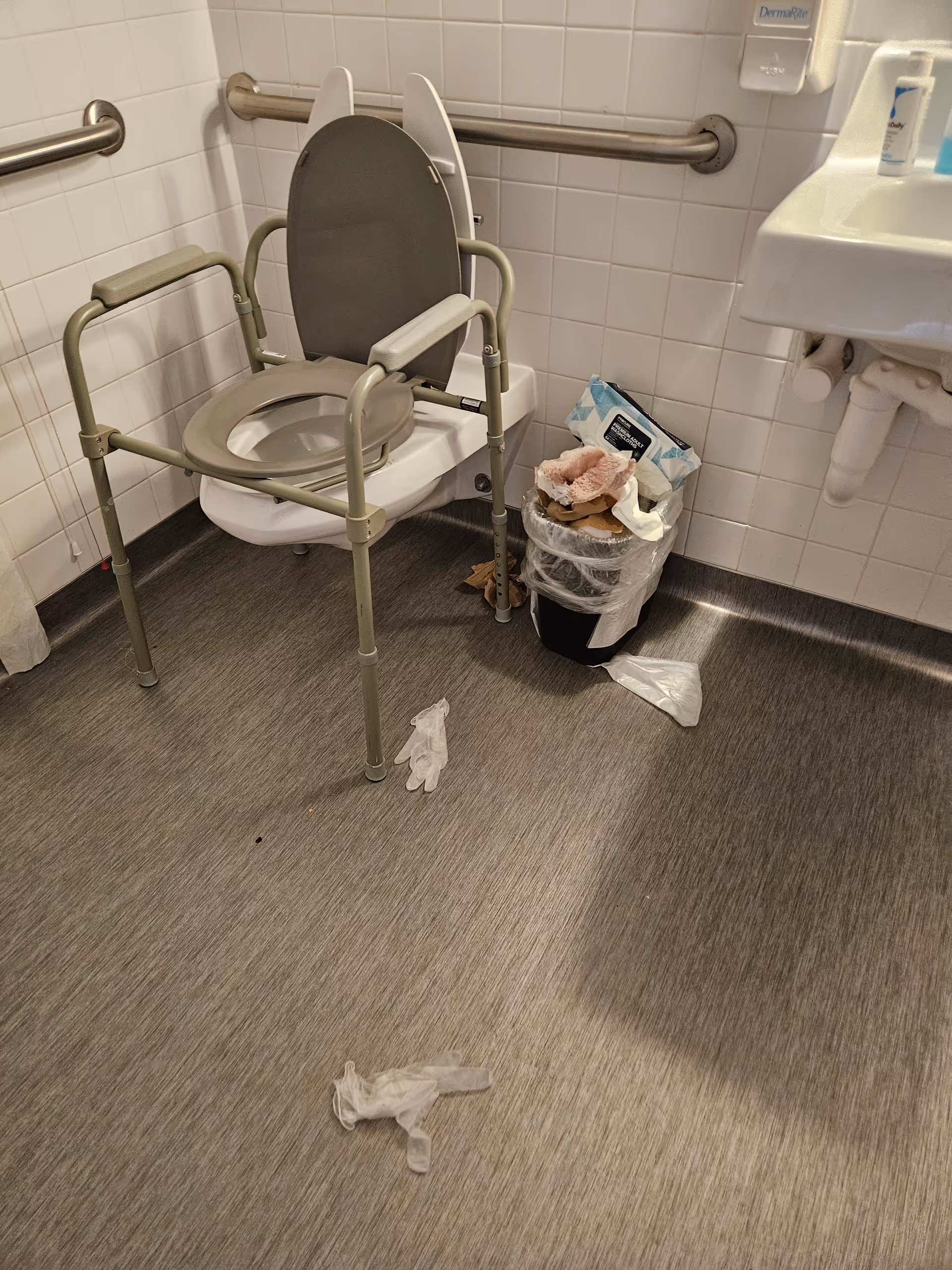 Accessible bathroom with a commode chair, sink, grab bars, and scattered trash.