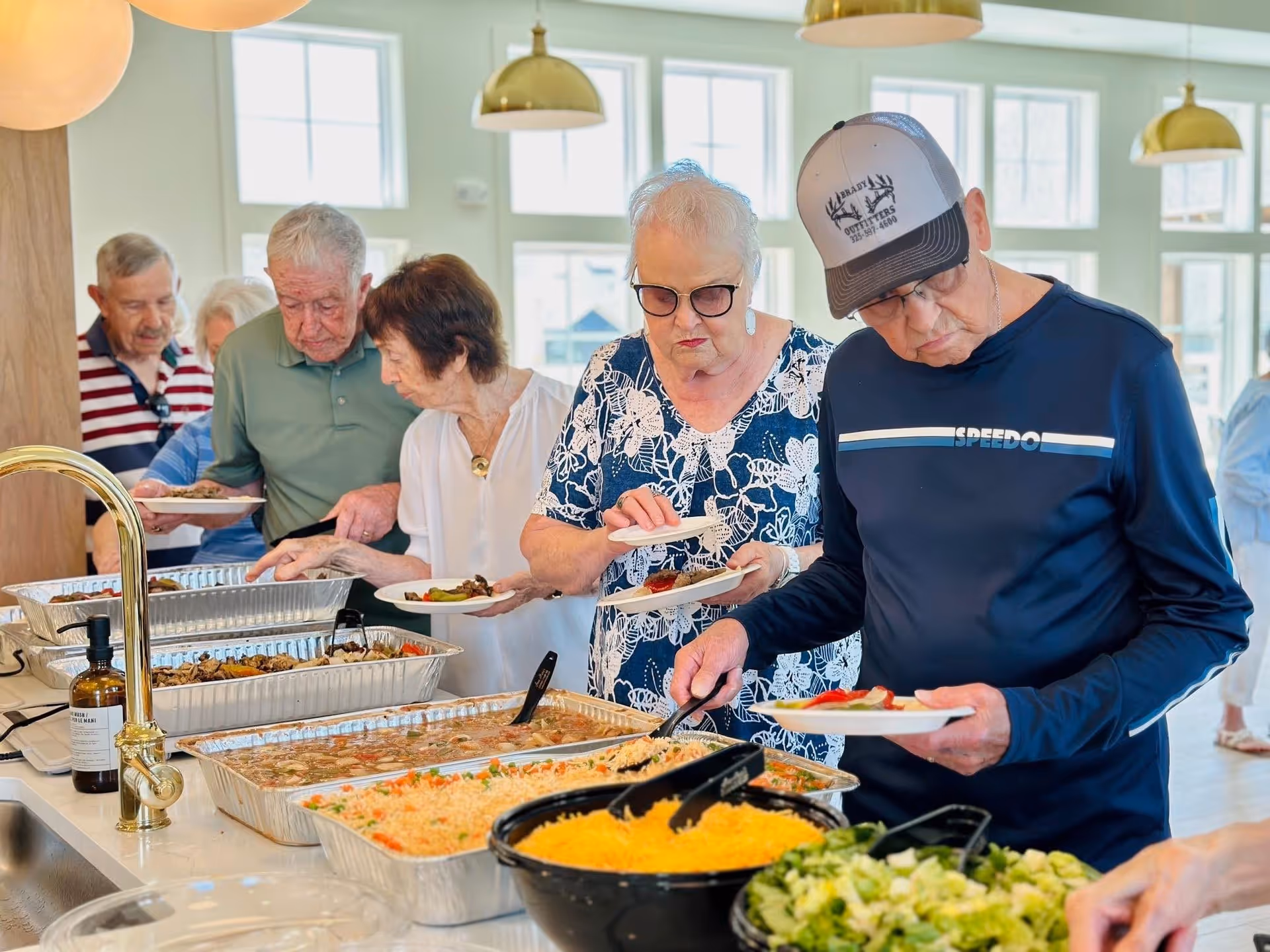 A group of elderly people serving themselves food from a buffet-style setup in a bright room with large windows and hanging pendant lights. The food includes rice, salad, and other dishes in large trays on a counter.