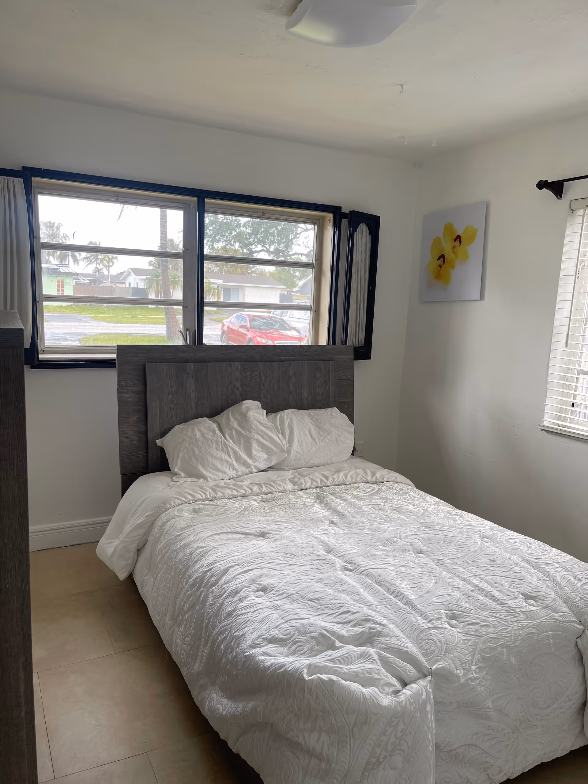 A neatly made bed in a small bedroom with windows and a wall painting.