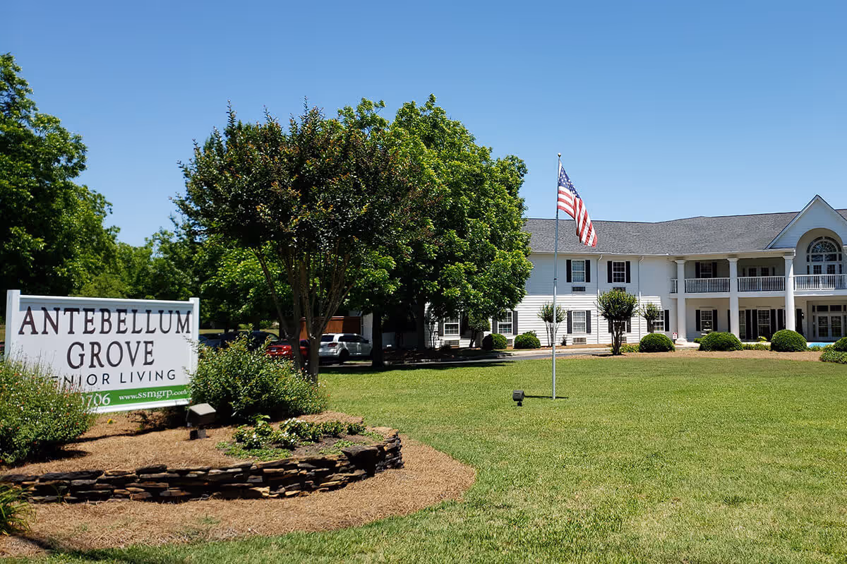 A two-story white senior living building with an American flag on the lawn and a sign reading 'Antebellum Grove Senior Living' in the foreground.