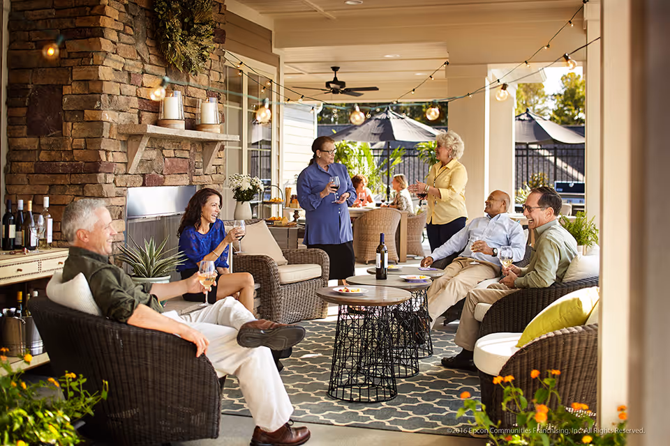 A group of six older adults socializing and enjoying drinks on a covered outdoor patio with wicker furniture, a stone fireplace, string lights, and a patterned rug. The setting is bright and inviting with plants and umbrellas visible in the background.