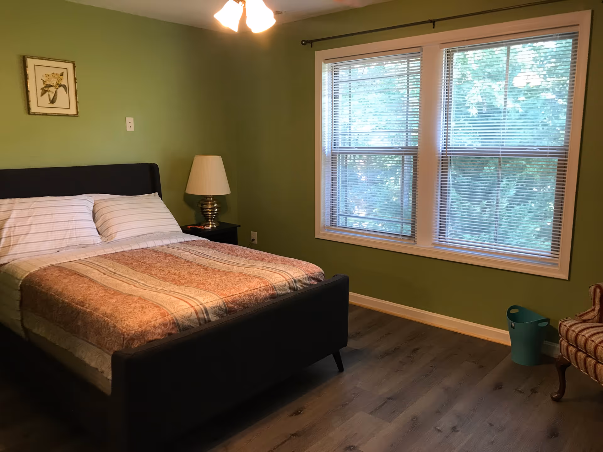 Bedroom with green walls, a made double bed, nightstand and lamp, large window with blinds, and wood-look flooring.