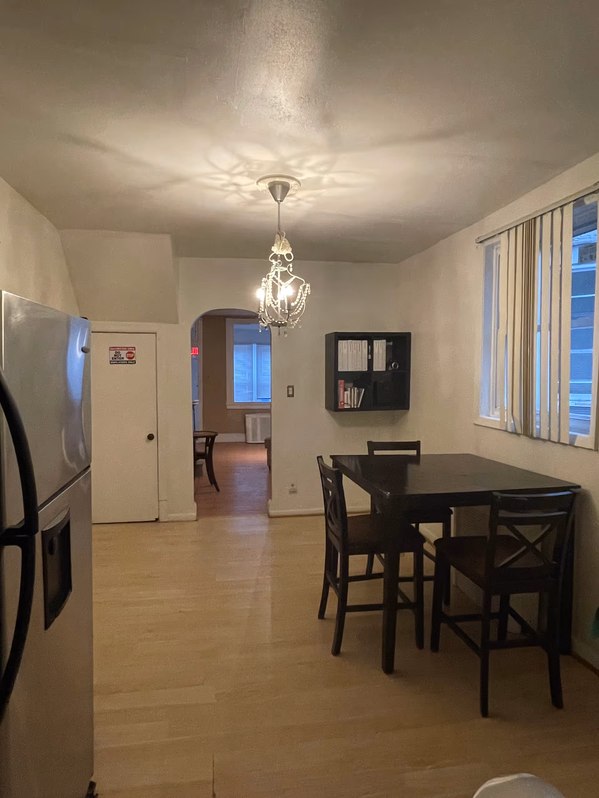 A dining area with a dark table and chairs, chandelier, and refrigerator along the wall.