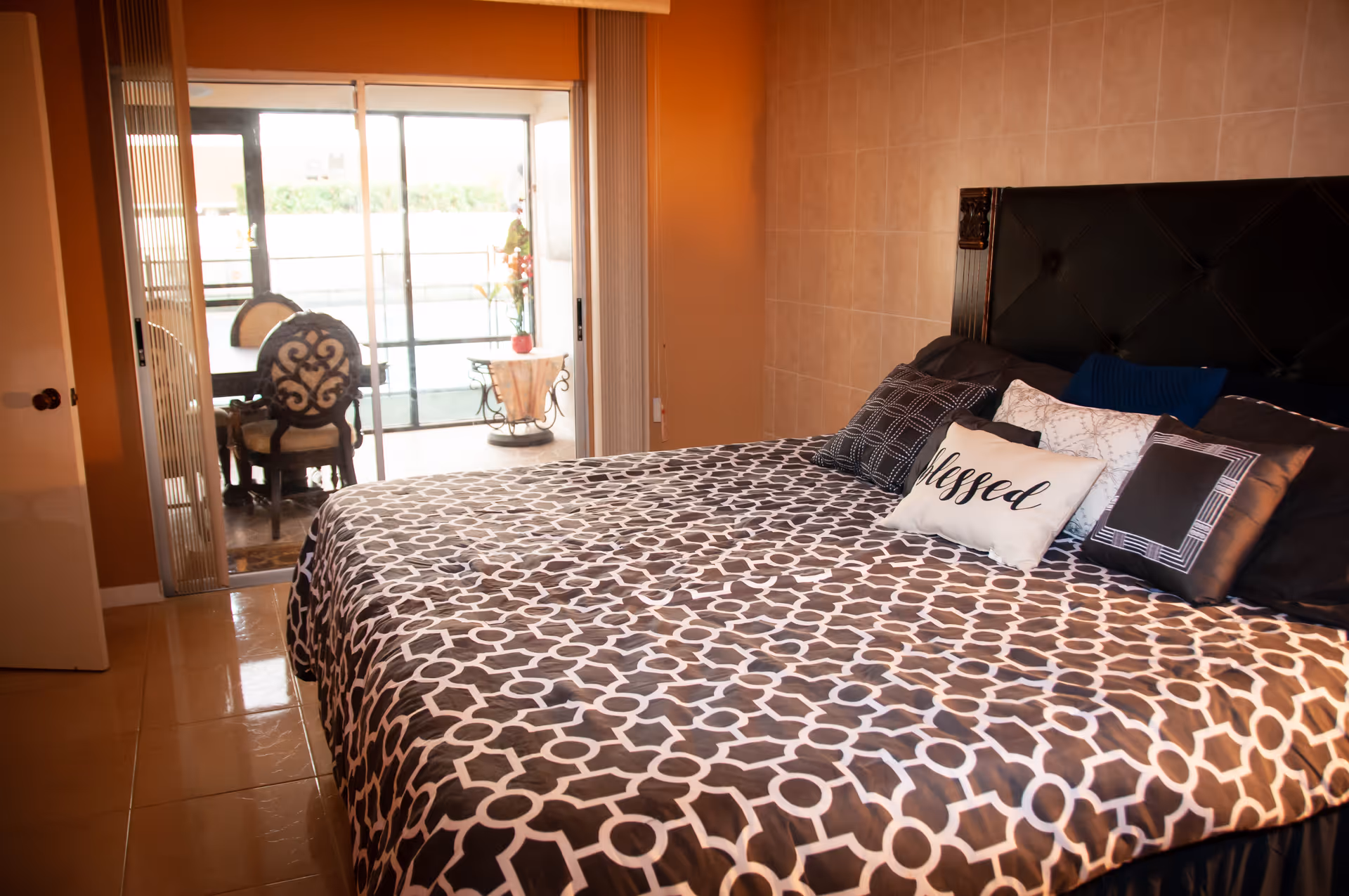 A bedroom with a large bed covered in a black and white patterned bedspread and multiple pillows, including one with the word 'blessed'. The room has tiled walls and floor, and a sliding glass door leading to a small outdoor patio area with a table and chairs.