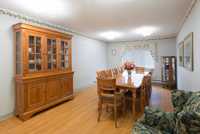 A dining room with a long wooden table surrounded by eight wooden chairs with patterned cushions. A vase with colorful flowers is placed in the center of the table. On the left side of the room, there is a large wooden cabinet with glass doors displaying dishes. On the right side, there is a floral patterned couch and two framed pictures on the wall. At the far end, a window with blinds and a decorative valance lets in natural light. The room has wooden flooring and a floral border along the top of the walls.