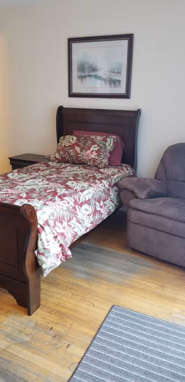 A bedroom with a single wooden bed featuring a floral patterned bedspread and matching pillow. Next to the bed is a dark wooden nightstand. A cushioned recliner chair is positioned beside the bed. Above the bed hangs a framed landscape painting. The floor is wooden with a gray striped rug partially visible.