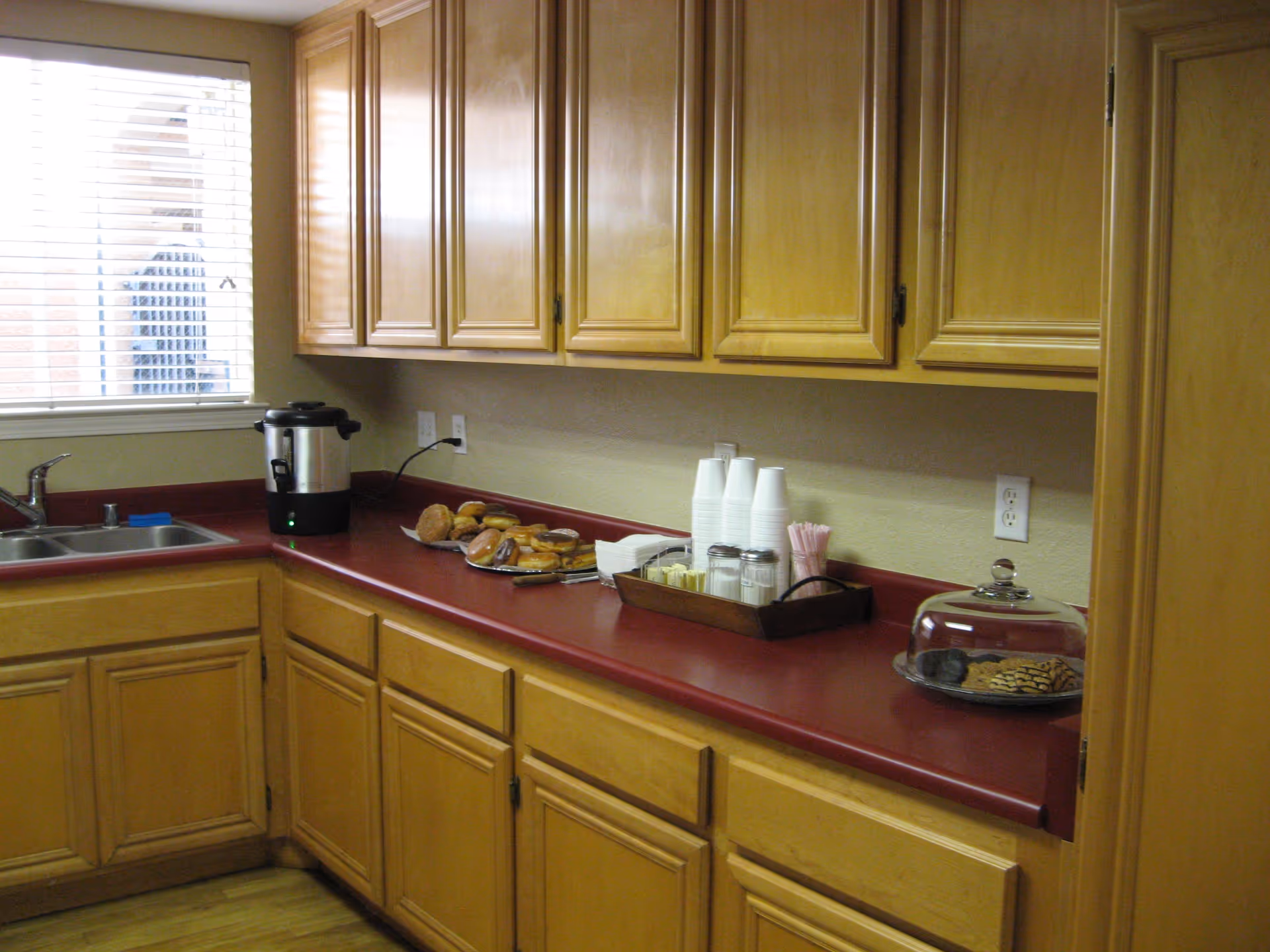 Kitchen area with wooden cabinets and a red countertop. On the counter, there is a coffee urn, a plate of assorted bagels and pastries, a tray with disposable cups, sugar, creamers, and stirrers, and a glass dome covering a plate of cookies. A window with blinds is visible above the sink.