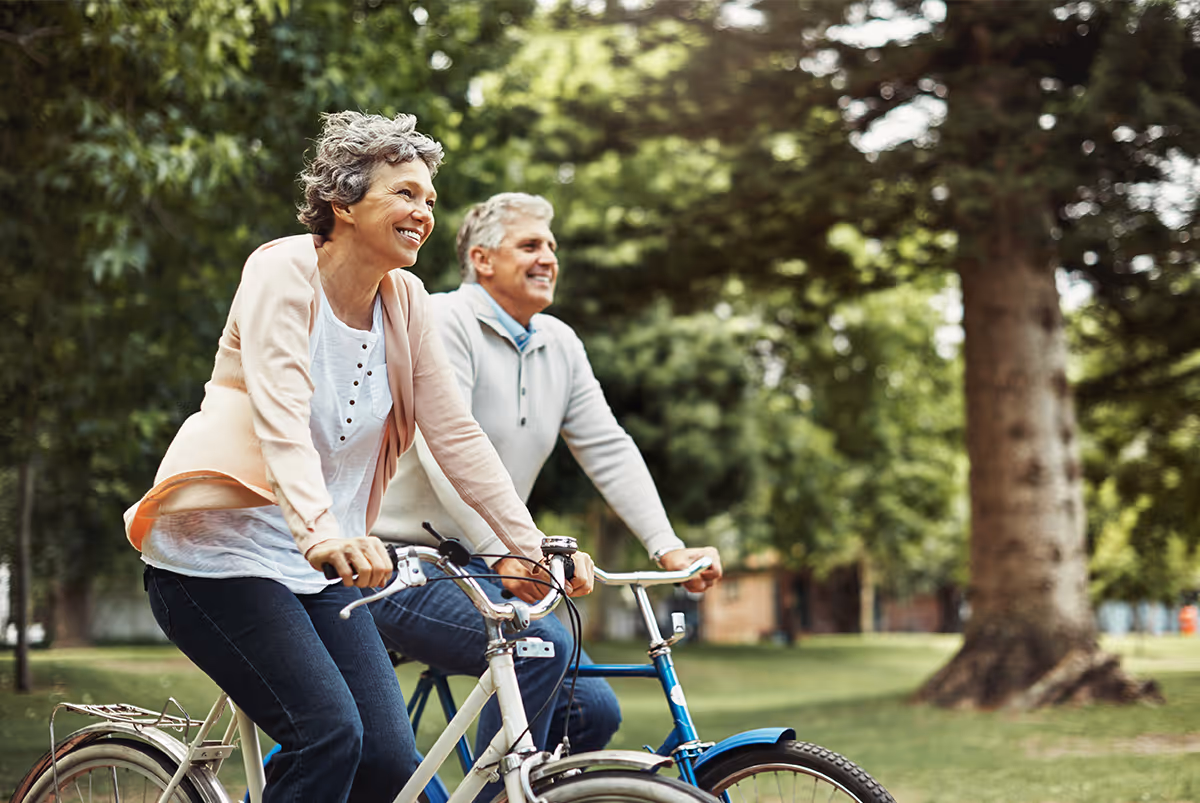 Two seniors ride bicycles together in a sunny outdoor park setting.