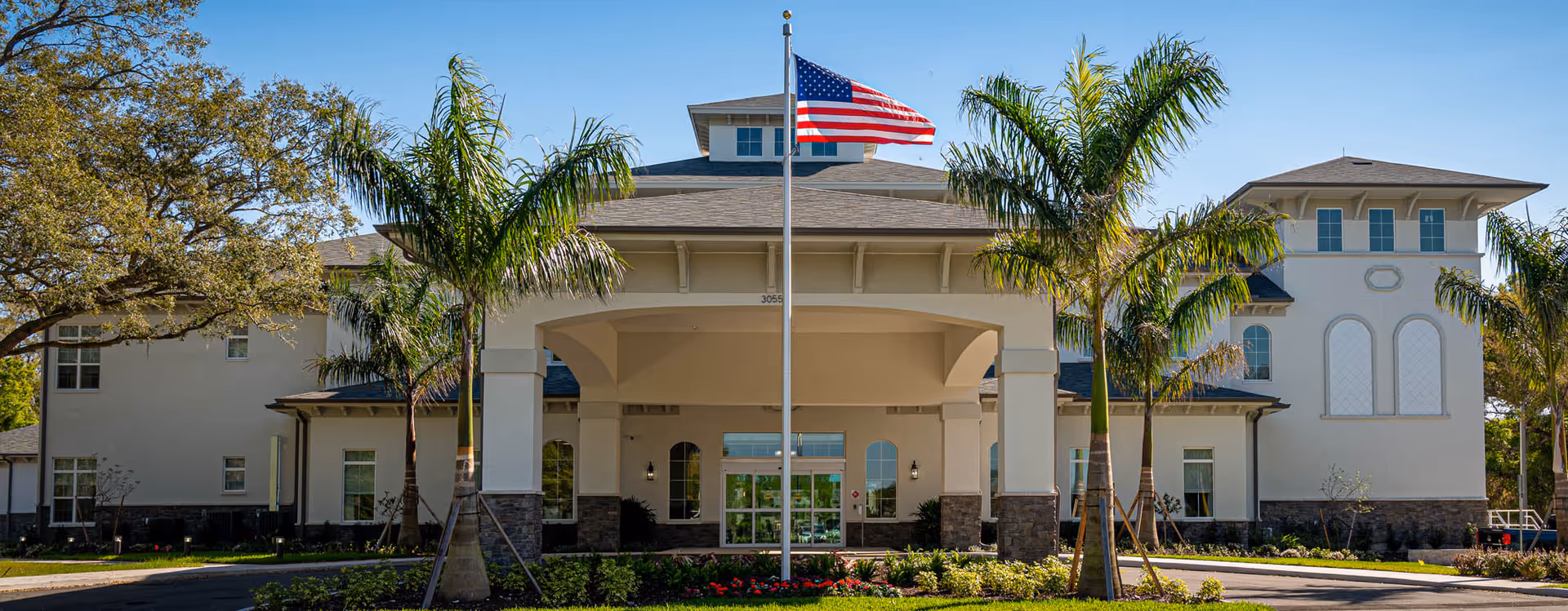 Front exterior view of a large, well-maintained building with a covered entrance, palm trees, and an American flag flying on a flagpole in front. The sky is clear and blue.