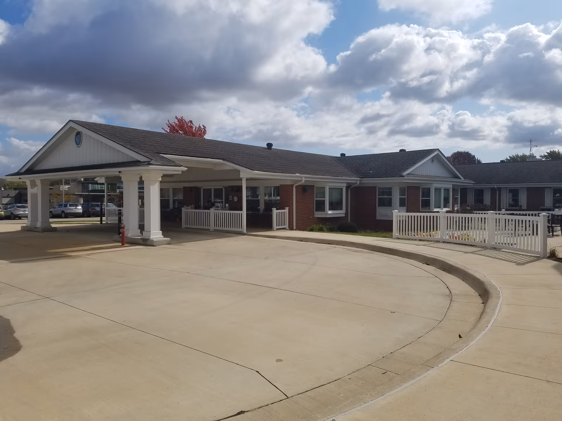 Exterior view of a single-story brick building with a covered entrance and white columns, surrounded by a concrete driveway and white fencing under a partly cloudy sky.