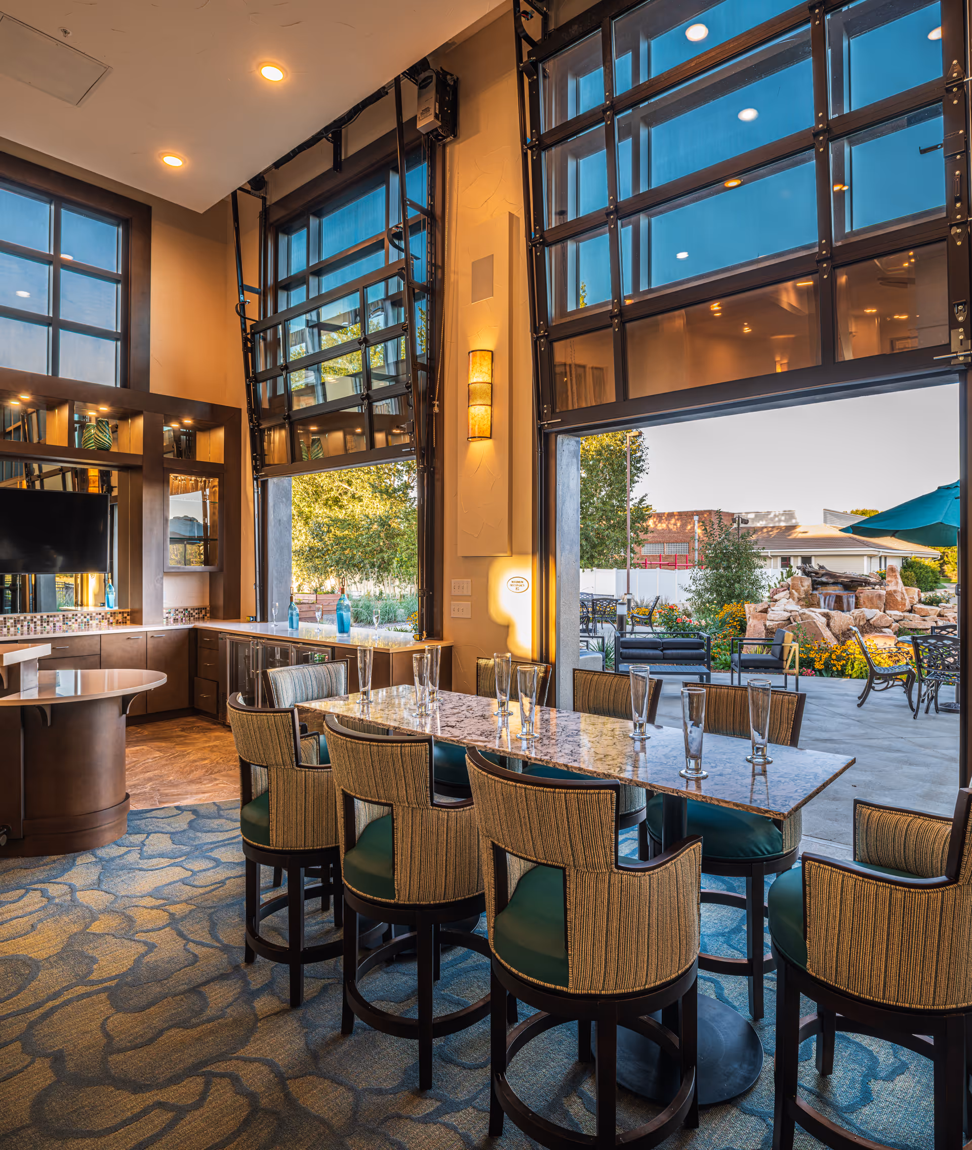 Interior view of a dining area with a long marble table surrounded by eight cushioned chairs. Large windows and glass garage-style doors are open, revealing an outdoor patio with seating, greenery, and a rock waterfall feature. The room has warm lighting, a patterned carpet, and a TV mounted on the wall.