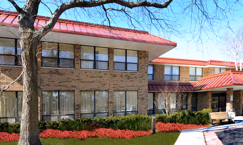 Exterior front view of a two-story brick senior living building with a red metal roof, rows of windows, landscaped flower beds and a bench.