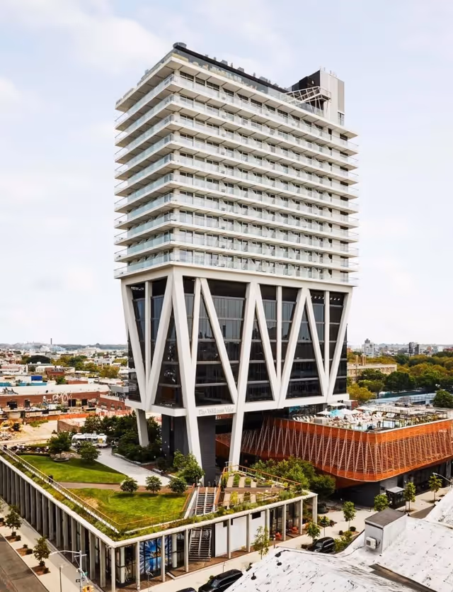 A tall modern building with diagonal white supports and glass facade, surrounded by greenery.