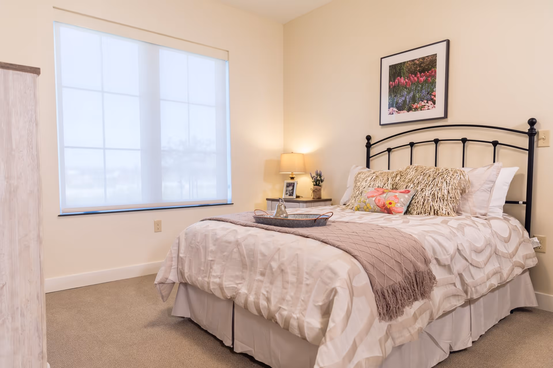 A cozy bedroom with a neatly made bed featuring a patterned beige and white comforter, decorative pillows, and a mauve throw blanket. A bedside table with a lamp, a small plant, and a framed photo is next to the bed. A large window with a white roller shade lets in natural light. A framed picture of flowers hangs on the wall above the bed.