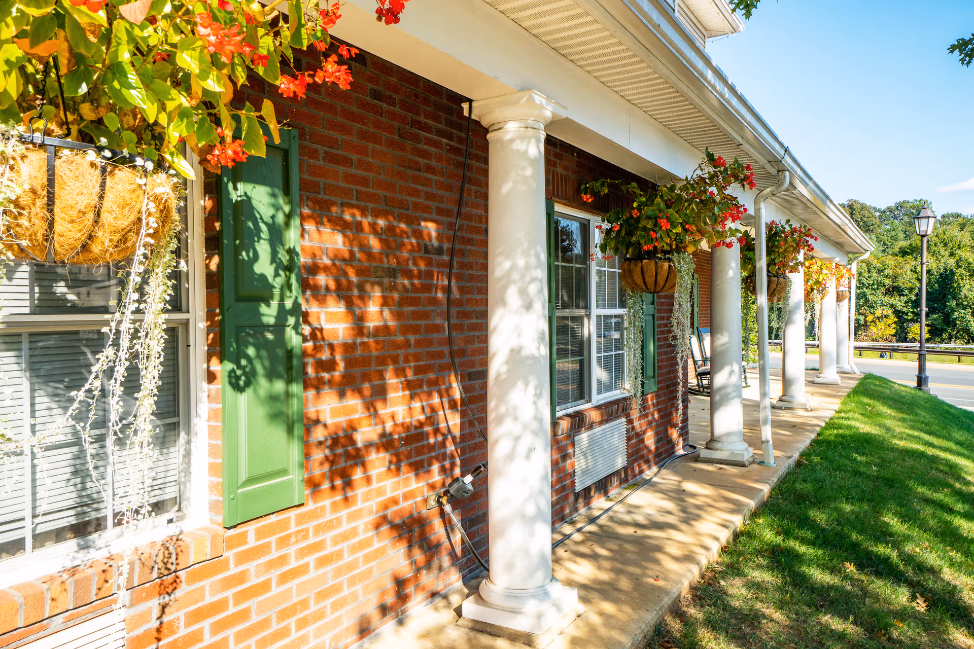 Brick building with green shutters, hanging flower baskets, and a white-columned porch along a sunny walkway.